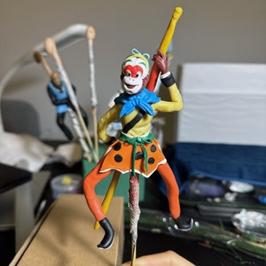 May include: A colourful clay figurine of a monkey wearing a yellow and black outfit with a blue scarf and holding a yellow stick. The monkey has a red face with white markings and a green crown.
