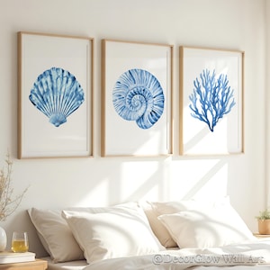 Puede incluir: Tres impresiones enmarcadas en acuarela con motivos marinos. Las impresiones muestran una concha azul, una concha de nautilus y coral sobre un fondo blanco. Los marcos son de madera clara y la obra de arte se exhibe sobre una cama con ropa de cama blanca.