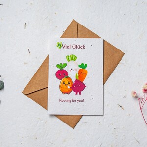 May include: A greeting card with a brown envelope. The card features a colorful illustration of cartoon vegetables with the text "Viel Glück" and "Rooting for you!". The background is a textured, off-white surface.