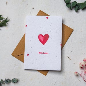 May include: A greeting card with a red watercolor heart and the words "With Love" on a white background. The card is on a brown envelope. Small red hearts are scattered around the card. The image is decorated with sprigs of greenery and flowers.