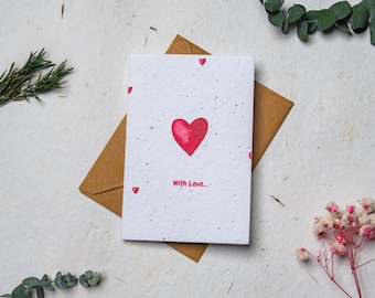 May include: A greeting card with a red watercolor heart and the words "With Love" on a white background. The card is on a brown envelope. Small red hearts are scattered around the card. The image is decorated with sprigs of greenery and flowers.