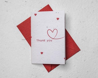 May include: A white card with the words "Thank you" in red, accompanied by a red heart design and small red hearts. The card rests on a red envelope, all set against a textured white background.