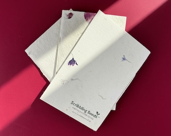 May include: A stack of handmade paper cards with embedded flower petals and small purple flower illustrations. The cards are arranged on a deep red surface, with the text "Scribbling Seeds" visible on one card.