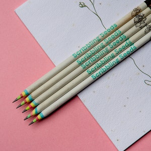 Könnte beinhalten: Fünf Holzbleistifte mit bunten Streifen und dem aufgedruckten Text "Scribbling Seeds". Die Stifte liegen auf einem weißen Blatt Papier mit einer kleinen Blumenzeichnung.