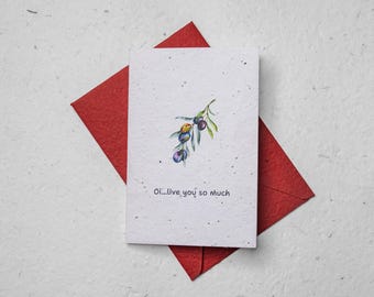 May include: A greeting card with a watercolor illustration of an olive branch with purple and green olives. The card is on a red envelope and has the text "Oi...live you so much". The card is on a textured white surface.