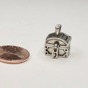 May include: A silver-colored treasure chest charm pendant, shown next to a US penny for size comparison. The charm has a hinged lid and detailed carvings.
