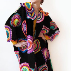 May include: A long, black crochet coat with large, colorful circular patterns in shades of pink, orange, yellow, green, and blue. The coat has a shawl collar and is worn with a matching head covering and green boots. The overall style is bohemian.