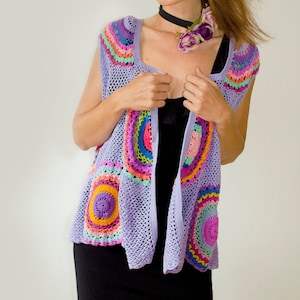 May include: A purple crochet vest with colorful circular patterns. The vest is sleeveless and has a loose fit. The model is wearing a black dress underneath.