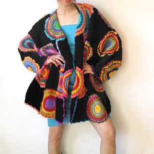 May include: A black crochet cardigan with colorful circular patterns. The cardigan has a loose fit and is open in the front. The model is wearing a blue dress and black boots.