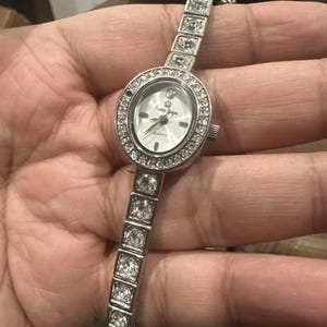 May include: Silver-toned quartz watch with a white face and a bezel and band adorned with clear crystals. The watch face displays the text "Prima Designs" and "QUARTZ". The band is composed of rectangular crystal accents.
