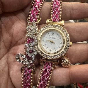 May include: A gold-toned wristwatch with a white face and crystal accents. The watch features a pink and gold band with the word "love" spelled out in crystals. The watch face has the text "Fah Teekay".