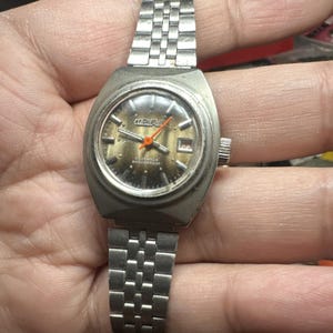 May include: Vintage silver-toned CORNAVY wristwatch with a rectangular face. The watch features a silver band, a gold-toned face, and orange hands. The watch face displays the brand name and the words "23 JEWELS SHOCKPROOF".