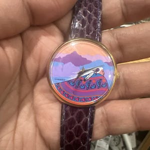 May include: A round wristwatch with a gold-tone bezel and a purple leather band. The watch face features a colorful illustration of a Native American figure with a mountain backdrop. The band is approximately 0.75 inches wide.