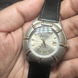 May include: A silver-toned wristwatch with gold-colored accents and a black leather band. The watch face has a textured design and displays the time. The word "OPEN" is visible on the watch.