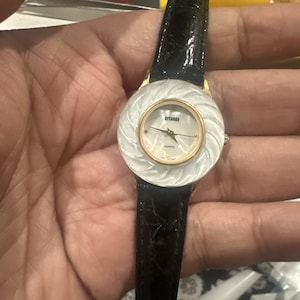May include: A gold-toned quartz watch with a white, textured face and a black leather band. The watch face has the brand name "GITANDE" and the word "QUARTZ" printed on it. The watch is a classic accessory.