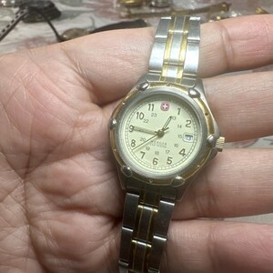 May include: A Wenger wristwatch with a two-tone silver and gold metal link band. The round watch face features a cream-colored dial with black Arabic numerals for 1-12 and 13-24 hour markers, a date window at the 3 o'clock position, and a red logo.
