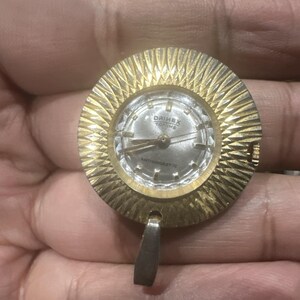 May include: A vintage gold-toned pendant watch with a textured, circular frame. The watch face is silver with the text "DRIMEX TOP TIME" and "ANTIMAGNETIC". A silver-toned bail hangs below the watch.