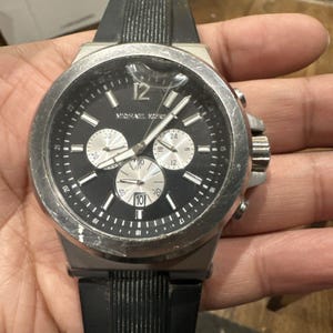 May include: A Michael Kors men's watch with a black face, silver bezel, and black rubber strap. The watch features three sub-dials and luminous hands. The watch face displays the brand name and has a date display.
