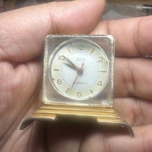 May include: A small square desk clock with a gold-toned base and a clear casing. The white circular clock face features gold-colored numerals at the 2, 4, 6, 8, 10, and 12 positions, with the text Hilton Jeweller and Swiss printed on the dial.
