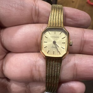 May include: A vintage gold-toned Wittnauer watch with a rectangular face and a woven band. The watch face is a light gold color with black hour markers and hands. The watch band is a matching gold color.