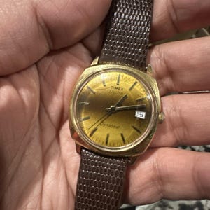 May include: A vintage Timex Electric Dynabeat watch with a gold-tone case and a brown leather strap. The watch face is gold with black hands and hour markers. The watch displays the day of the week.