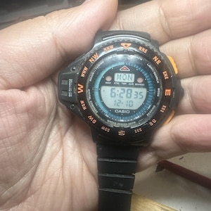May include: A black Casio digital watch with a round face featuring a compass bezel with orange cardinal directions. The display shows the time 6:28, the date 12-10, and the day MON. It includes a black resin strap and a bearing sensor label.