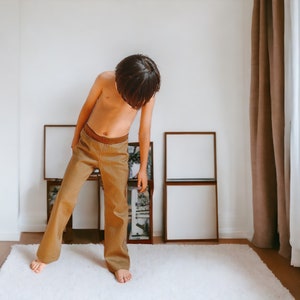 Op de afbeelding: Een jong persoon in een bruine broek staat in een kamer met witte muren en een tapijt. Er hangen verschillende ingelijste foto's aan de muur achter hen.
