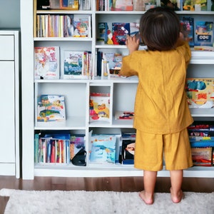 May include: A young child in a yellow outfit reaches for a book on a white bookshelf filled with colorful books.