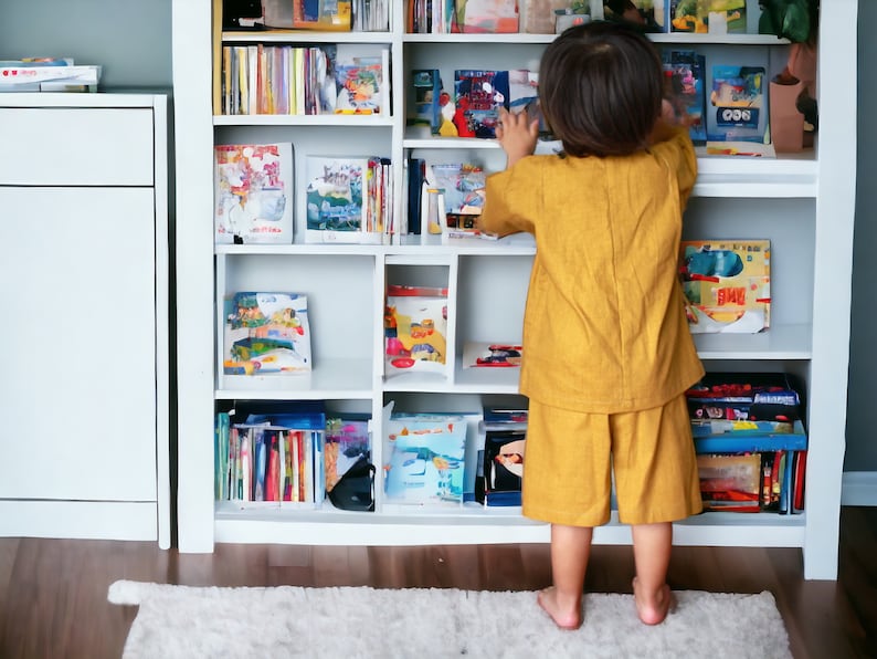 May include: A young child in a yellow outfit reaches for a book on a white bookshelf filled with colorful books.
