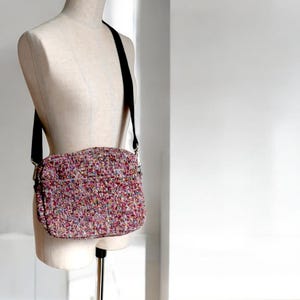 May include: A multi-colored crossbody bag with a black strap is displayed on a mannequin. The bag has a textured, woven appearance with a mix of pink, purple, and other colors. The strap is adjustable and appears to be made of a durable material.