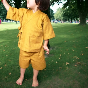 May include: A young child wearing a yellow linen kimono-style outfit in a park setting.