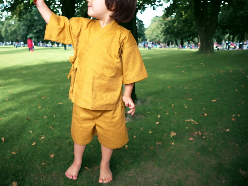 May include: A young child wearing a yellow linen kimono-style outfit in a park setting.