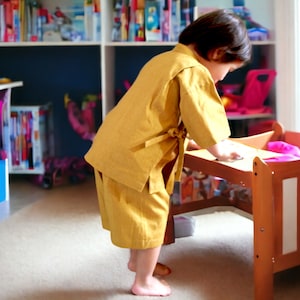 May include: A young child wearing a yellow robe-like garment stands near a wooden doll bed. The child is reaching towards the bed with their right hand.