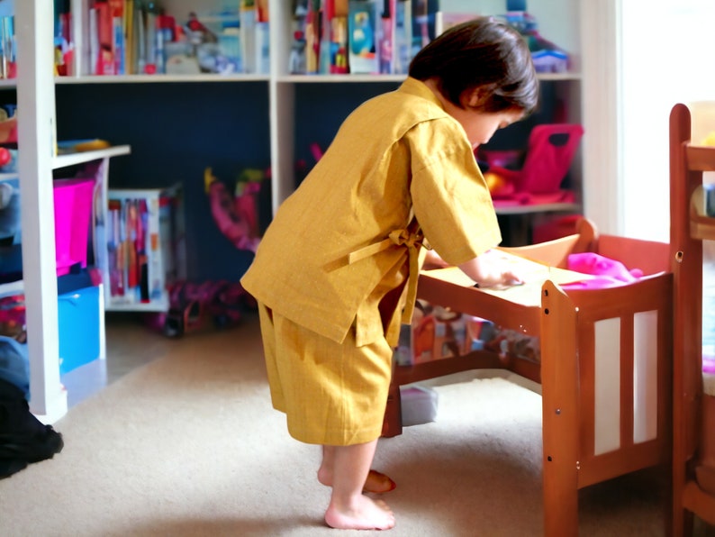 May include: A young child wearing a yellow robe-like garment stands near a wooden doll bed. The child is reaching towards the bed with their right hand.