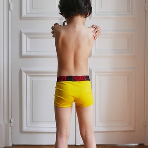 May include: Bright yellow boxer briefs featuring a black elastic waistband with the word PORT printed in bold red capital letters. The garment is constructed from a ribbed fabric and sits at the waist, providing a snug fit for daily wear.