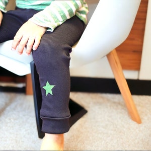 May include: A child wearing dark gray ribbed leggings with a green star on one leg. The child is sitting on a white chair with wooden legs.