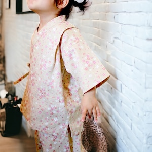 May include: A young child wearing a light brown and pink patterned kimono with a white background. The kimono has a floral design and a long sleeve. The child is standing in front of a white brick wall.