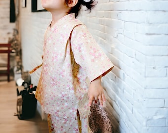 May include: A young child wearing a light brown and pink floral print kimono-style top and matching pants. The top has a long sleeve and a collar. The child is standing near a white brick wall.