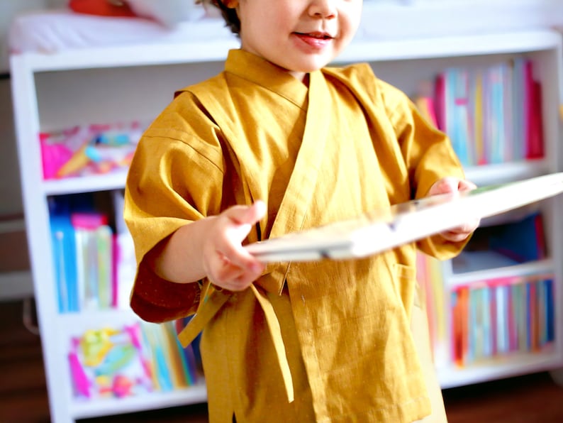 May include: A young person wearing a yellow kimono-style robe holds a book open in front of them. The book is white with a colorful cover. The person is standing in front of a bookshelf filled with colorful books.