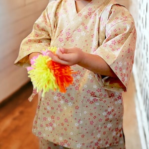 May include: A child wearing a light brown and pink patterned kimono holds a colorful yarn ball in their hands. The yarn ball is made of yellow, orange, and pink yarn.