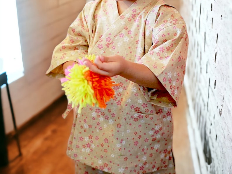 May include: A child wearing a light brown and pink patterned kimono holds a colorful yarn ball in their hands. The yarn ball is made of yellow, orange, and pink yarn.