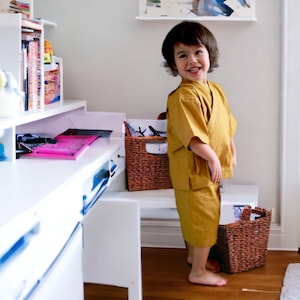 May include: A young child wearing a yellow kimono-style outfit stands in front of a white desk with a pink folder on top. The child is smiling and looking at the camera. There are two wicker baskets on the floor.