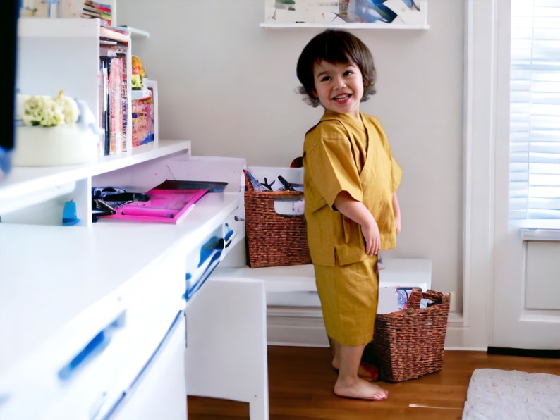 May include: A young child wearing a yellow kimono-style outfit stands in front of a white desk with a pink folder on top. The child is smiling and looking at the camera. There are two wicker baskets on the floor.