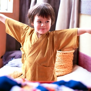 May include: A young person wearing a yellow robe with long sleeves, standing with arms outstretched, smiling at the camera.