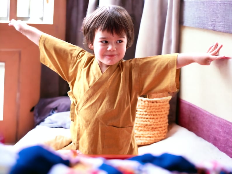 May include: A young person wearing a yellow robe with long sleeves, standing with arms outstretched, smiling at the camera.