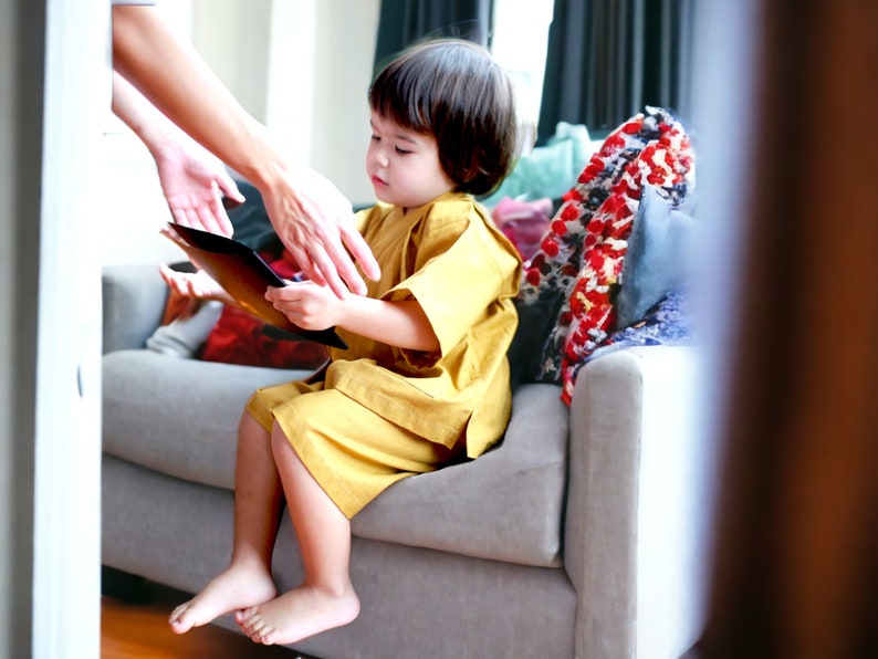 May include: A young child in a yellow dress sits on a gray couch and holds a black tablet. An adult's hand reaches towards the child.