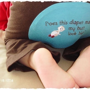 Puede incluir: Pantalones de bebé marrones con un parche azul claro con el texto "Does this diaper make my butt look big?" y un bebé de dibujos animados. El bebé lleva una camisa roja.