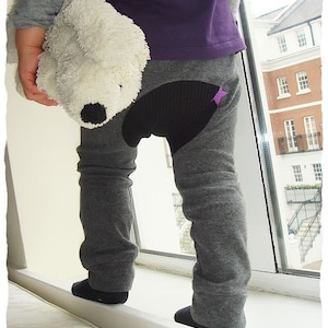 Pode incluir: Uma criança vestindo calças cinzas com um remendo de estrela preta e roxa no joelho esquerdo. A criança está segurando um animal de pelúcia branco.