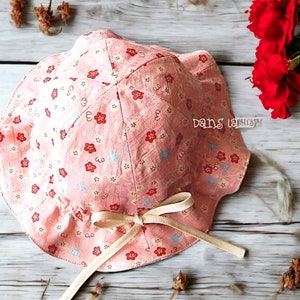 Può includere: Un cappello da sole in tessuto rosa con un nastro bianco e una stampa floreale. Il cappello ha un bordo arricciato ed è legato con un fiocco.