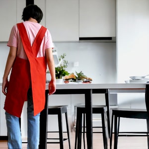 May include: A person wearing a red apron and blue jeans stands in a kitchen with a white countertop and black bar stools.